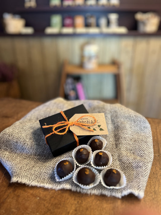 A gift set of chocolate truffles and a small black box tied with orange ribbon and a paper tag reading 'chocolate', arranged on a textured gray cloth on a wooden table with a blurred indoor background.